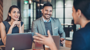 Corporate visibility; man and woman smiliing, sitting next to each other listening to woman sitting across speaking to them