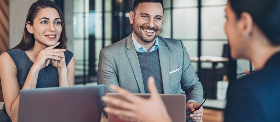 Corporate visibility; man and woman smiliing, sitting next to each other listening to woman sitting across speaking to them