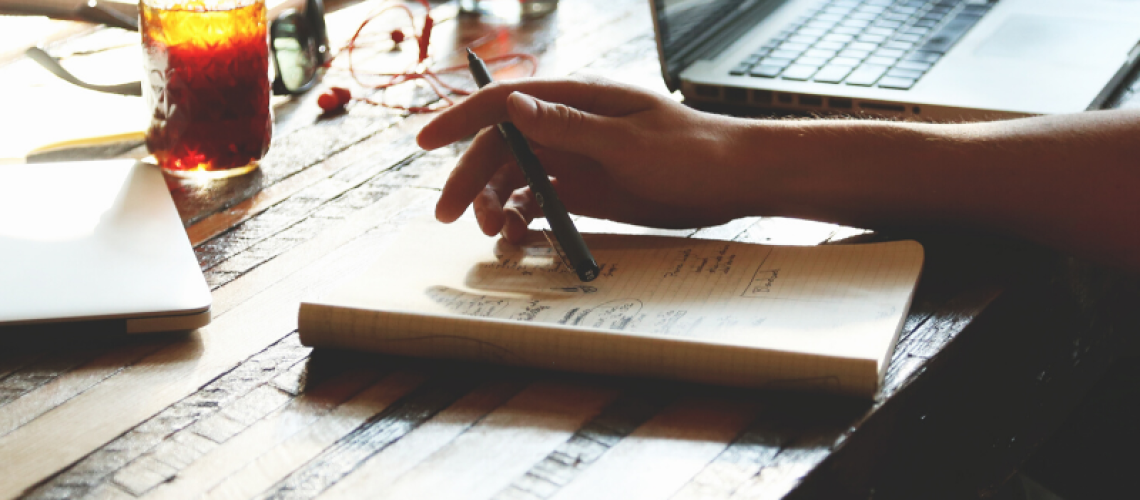 man's hand holding a pen on top of a notebook, lap top beside him